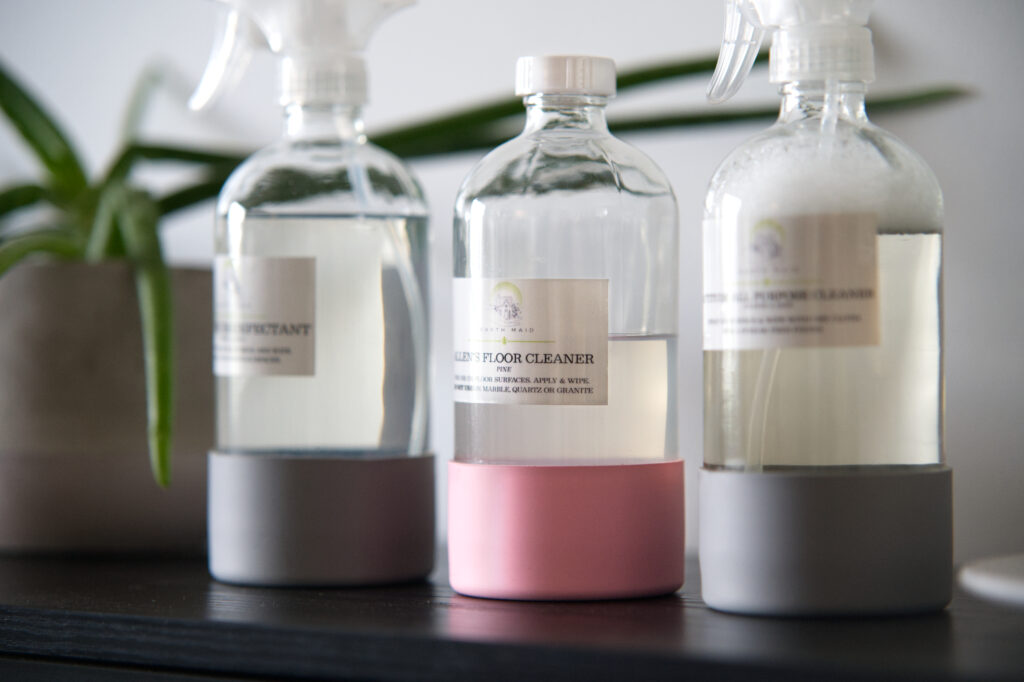 Three clear cleaning bottles with white labels and pastel bases (pink and gray) lined up on a dark surface; center bottle labeled floor cleaner.