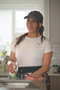 Smiling woman in a white Earth Maid shirt and dark baseball cap stands at a kitchen counter, holding a spray bottle and a pink cloth.