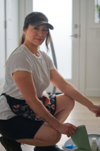 Woman kneeling indoors, wearing a cap and white shirt, holding a green glove beside a bucket of cleaning supplies in a bright room.