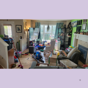 Cluttered living room with toys, furniture, and storage bins scattered across the floor; large window with blue curtains behind a chair and bookshelf units to the right.
