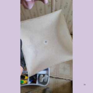 Beige textured cushion with a central circular hole, held by a gloved hand against a tiled floor background.
