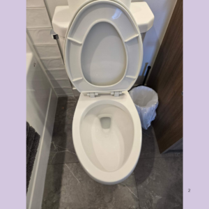 White toilet with the lid up in a small bathroom; dark gray tiled floor and a trash can to the right.