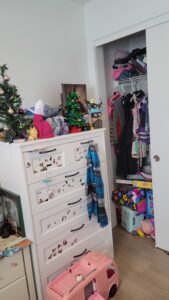 Cluttered children's bedroom with a white dresser covered in stickers and decor, a small Christmas tree, and an open closet full of clothes and shelves.
