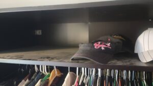 Open closet shelf with two caps resting on top and colorful shirts hanging below on a rod.