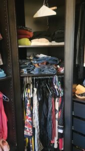 Open closet with folded sweaters on top shelf, jeans and belts on middle shelf, and hanging dresses on the lower rod all organized in a dark wardrobe.
