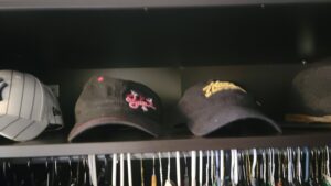 Shelf with several baseball caps in a closet: gray striped cap, a black cap with pink floral embroidery, and a dark cap with yellow text visible on the right.