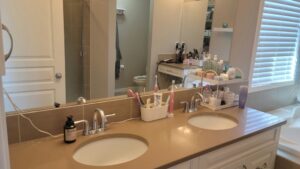 Bathroom vanity with double sink and beige countertop; cluttered with toiletries, hair tools, and a mirror with window blinds in the background.