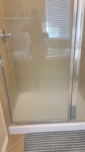 Glass shower door in a tiled bathroom; reflection shows a person taking a photo, window with blinds, a white tub, and a gray striped bath mat on the floor.