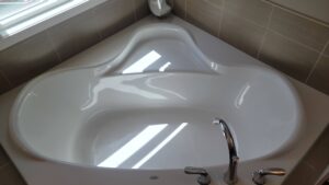 Corner white bathtub with chrome faucets in a tiled bathroom near a window, showing a curved built-in design.