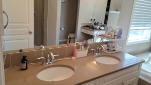 Double-sink bathroom vanity with makeup organizer, hair tools, and bottles on the counter; window with blinds in the background.