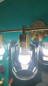 Close-up of a modern bathroom vanity light with exposed bulbs inside glass shades and brushed metal fittings against a teal wall.