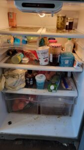 Open refrigerator with cluttered shelves: jars, bottles, yogurt cup, and packaged foods scattered inside.