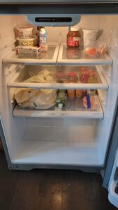 Open refrigerator with assorted groceries on three shelves: Nutella jar, protein drink, yogurts on top; juice bottles and condiments on lower shelves; plastic bag on bottom shelf.