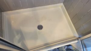 Empty shower stall with light gray tiled walls, a round metal drain in the center, and a slim threshold.