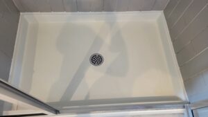 White rectangular shower base with circular drain in the center, grey tile walls surrounding it, and a glass door frame at the bottom edge
