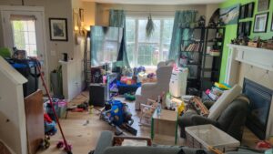 Messy living room filled with children's toys, clothing, and clutter strewn across the floor and furniture.
