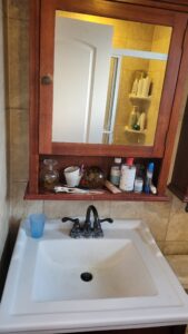 Bathroom vanity with a wooden framed mirror cabinet; cluttered shelf holds bottles, cups and grooming items above a white sink with a dark faucet.