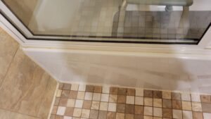 Shower door with a white frame next to beige tiled floor; a yellowish stain along the seal shows wear or corrosion.