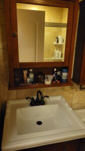 Wood-framed bathroom mirror cabinet with a shelf holding bottles and grooming items above a white sink.