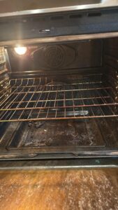 Oven interior with a light on, dirty bottom and a metal rack pulled out, showing grease and debris inside.